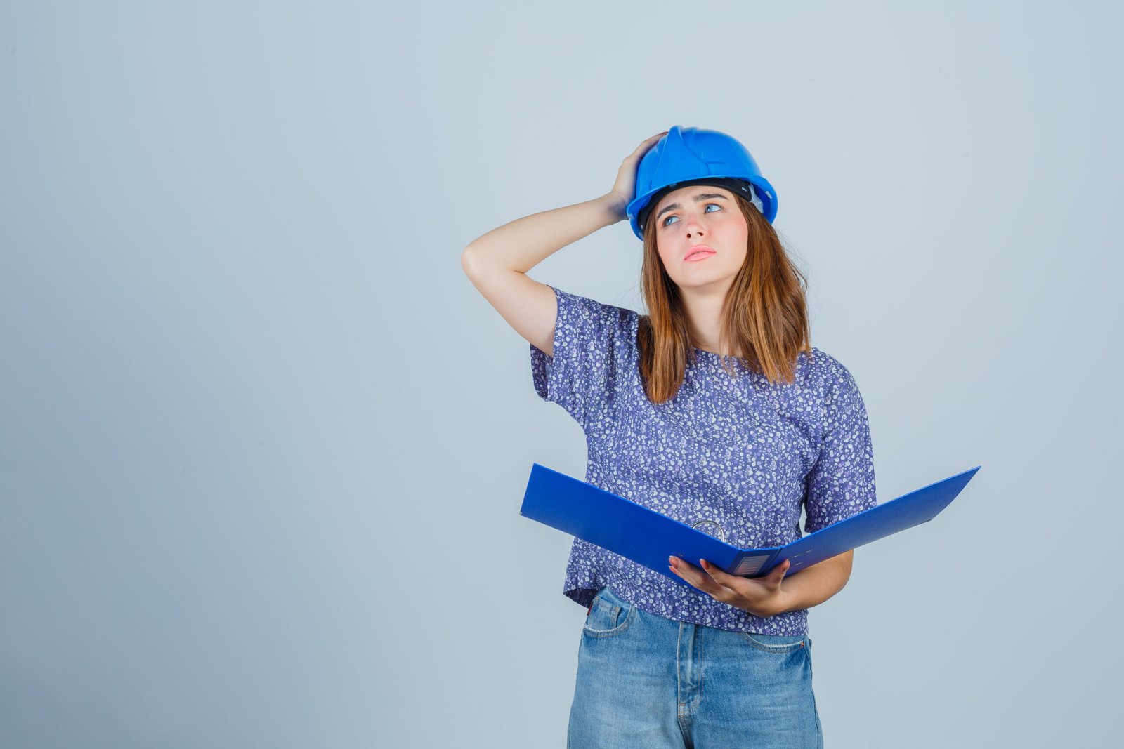 young girl holding folder, keeping hand on head in blouse, jeans, helmet and looking pensive. front view.