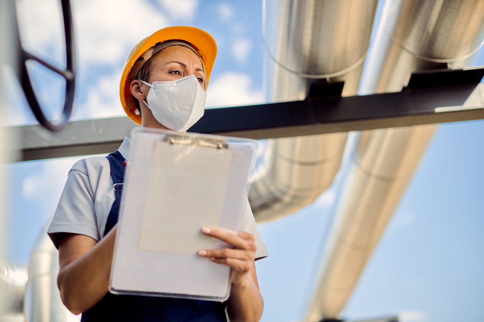 Low angle view of female civil engineer with face mask taking notes while inspecting project development at construction site